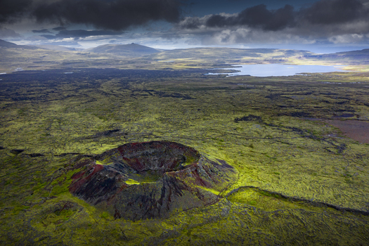 Aerial view of a volcanic crater surrounded by green mossy lava fields and a distant lake under a dramatic cloudy sky in Iceland.