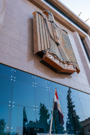 View of an Egyptian flag flying beneath a building with a large emblem against a blue glass facade a contrast of heritage and modernity Zamalek Cairo Governorate Egypt. View of an Egyptian flag flying beneath a building with a large emblem against a blue glass facade a contrast of heritage and modernity Zamalek Cairo Governorate Egypt.