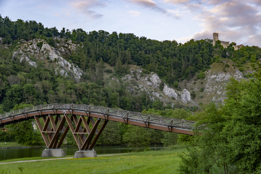 View of the wooden Tatzelwurm bridge crossing the Altmuhl river with Randeck Castle perched on a rocky hill surrounded by lush green forest in Essing Bayern Germany.