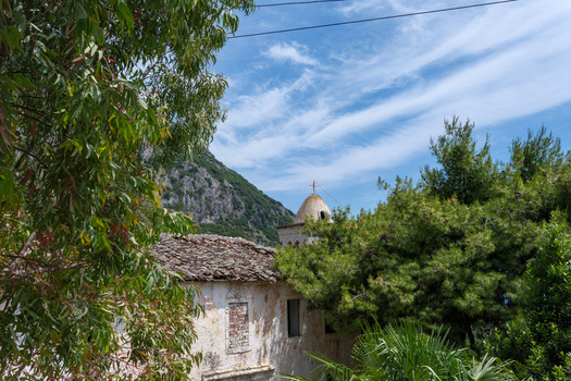 View of an old stone house with a traditional tiled roof and a church dome nestled among lush green trees and a rocky mountain under a blue sky Qeparo Fshat Vlore County Albania. View of an old stone house with a traditional tiled roof and a church dome nestled among lush green trees and a rocky mountain under a blue sky Qeparo Fshat Vlore County Albania.