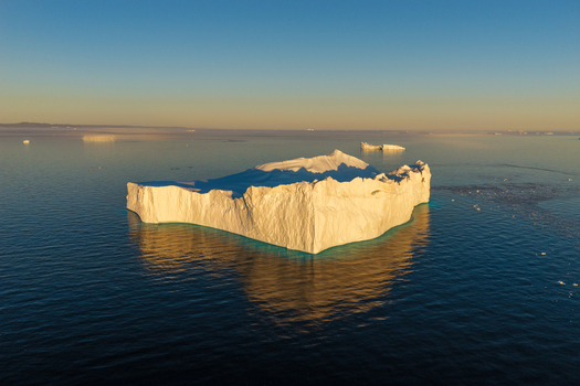 Aerial view of a massive iceberg floating in the calm deep blue waters of the Arctic illuminated by the warm glow of the golden hour in Greenland. Aerial view of a massive iceberg floating in the calm deep blue waters of the Arctic illuminated by the warm glow of the golden hour in Greenland.