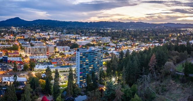 Aerial view of the city of Eugene at twilight featuring illuminated buildings dense surrounding forests and a colorful sunset sky over the horizon in Eugene Oregon United States. Aerial view of the city of Eugene at twilight featuring illuminated buildings dense surrounding forests and a colorful sunset sky over the horizon in Eugene Oregon United States.