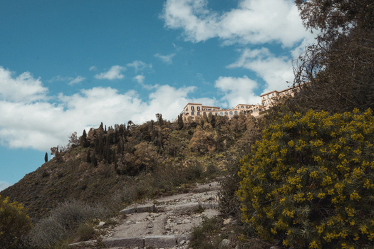 View of ancient stone stairs climbing toward a hilltop village amidst lush green trees against a backdrop of blue sky and white clouds Taormina Sicily Italy.