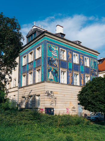 View of Warsaw Old Town building with blue and turquoise mosaic tiles depicting figures surrounded by trees under a clear blue sky in Warsaw Masovian Voivodeship Poland. View of Warsaw Old Town building with blue and turquoise mosaic tiles depicting figures surrounded by trees under a clear blue sky in Warsaw Masovian Voivodeship Poland.