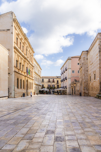 View of sun drenched piazza with elegant buildings casting shadows on the stone pavement creating a vibrant scene Ortigia Sicily Italy.
