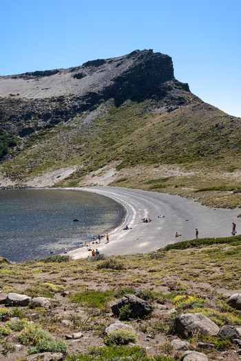 View of Volcan Batea Mahuida crater lake with dark volcanic sand beach clear blue sky and people relaxing on the shore in Alumine Neuquen Argentina. View of Volcan Batea Mahuida crater lake with dark volcanic sand beach clear blue sky and people relaxing on the shore in Alumine Neuquen Argentina.
