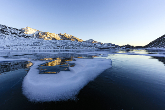 View of a frozen alpine lake with snow covered mountain peaks reflecting in the icy water under a clear blue sky in Madesimo Lombardy Italy. View of a frozen alpine lake with snow covered mountain peaks reflecting in the icy water under a clear blue sky in Madesimo Lombardy Italy.