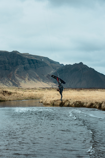 View of a kiteboarder performing a jump over a river with dramatic mountains and dry grassy banks under an overcast sky in Borgarbyggd Iceland. View of a kiteboarder performing a jump over a river with dramatic mountains and dry grassy banks under an overcast sky in Borgarbyggd Iceland.