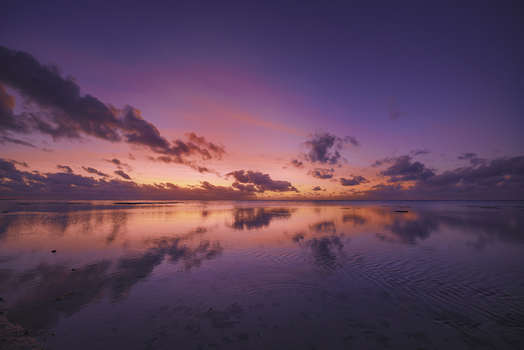 View of Aitutaki Lagoon at sunset with vibrant purple and orange clouds reflecting on the calm water surface in Aitutaki Cook Islands.