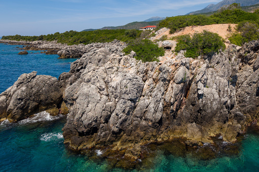 Aerial view of Aquarium Beach with rugged limestone cliffs turquoise sea water and a historic concrete bunker nestled in green Mediterranean scrub Jala Vlore County Albania.