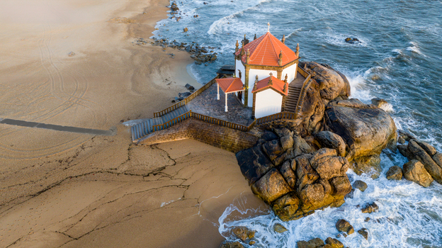 Aerial view of Capela do Senhor da Pedra on a rocky outcrop surrounded by sandy beach and crashing waves in Miramar Porto Portugal.