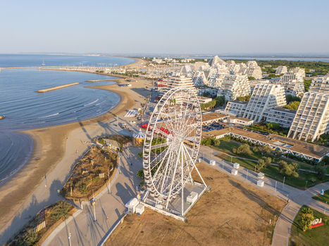 Aerial view of a large Ferris wheel situated on the sandy beach near the distinctive pyramid shaped architecture of La Grande Motte Occitanie France.