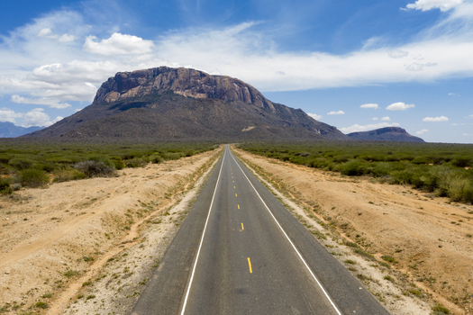 Aerial view of a long straight road stretching through the arid landscape towards a prominent volcanic mountain in Marsabit Marsabit County Kenya. Aerial view of a long straight road stretching through the arid landscape towards a prominent volcanic mountain in Marsabit Marsabit County Kenya.