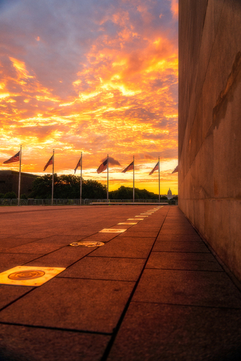 View of the base of the Washington Monument with American flags waving against an orange sunset sky and the distant Capitol building in Washington District of Columbia United States. View of the base of the Washington Monument with American flags waving against an orange sunset sky and the distant Capitol building in Washington District of Columbia United States.