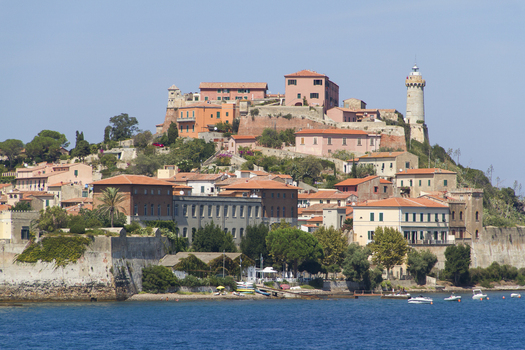 View of the pastel colored buildings cascade down the hillside towards the deep blue sea crowned by a lighthouse under a clear sky Portoferraio Tuscany Italy. View of the pastel colored buildings cascade down the hillside towards the deep blue sea crowned by a lighthouse under a clear sky Portoferraio Tuscany Italy.