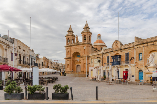 View of the Parish Church casting a warm hue over the quiet square lined with quaint cafes and buildings under an expansive sky Marsaxlokk Malta. View of the Parish Church casting a warm hue over the quiet square lined with quaint cafes and buildings under an expansive sky Marsaxlokk Malta.