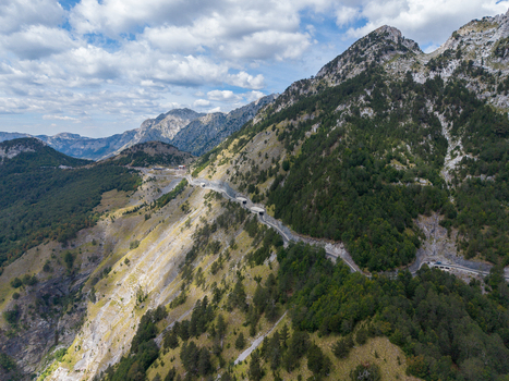 Aerial view of a winding mountain road through the rugged terrain of the Albanian Alps with lush green forests and rocky slopes under a bright sky in Shkoder Shkoder County Albania. Aerial view of a winding mountain road through the rugged terrain of the Albanian Alps with lush green forests and rocky slopes under a bright sky in Shkoder Shkoder County Albania.