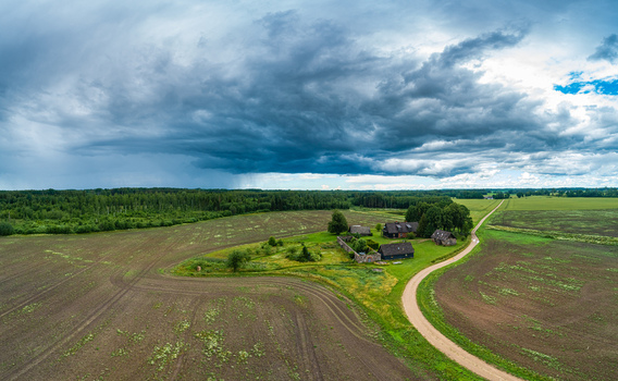 Aerial view of a rural farmstead with traditional wooden buildings surrounded by agricultural fields and a winding dirt road under a dramatic stormy sky in Latvia. Aerial view of a rural farmstead with traditional wooden buildings surrounded by agricultural fields and a winding dirt road under a dramatic stormy sky in Latvia.