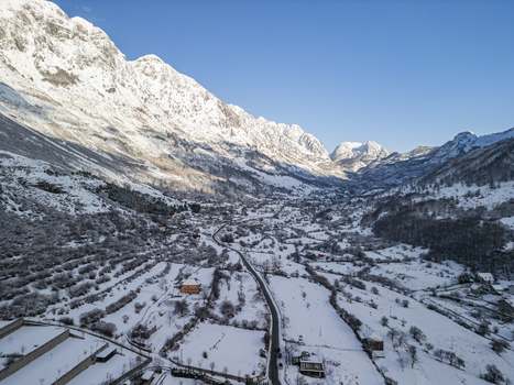Aerial view of a snow covered mountain valley in winter with a winding road scattered houses and rugged peaks under a clear blue sky in Boge Shkoder County Albania. Aerial view of a snow covered mountain valley in winter with a winding road scattered houses and rugged peaks under a clear blue sky in Boge Shkoder County Albania.