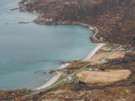 Aerial view of Senja island coastline featuring a winding road small houses and autumn colored vegetation along the blue sea in Botnhamn Troms Norway.