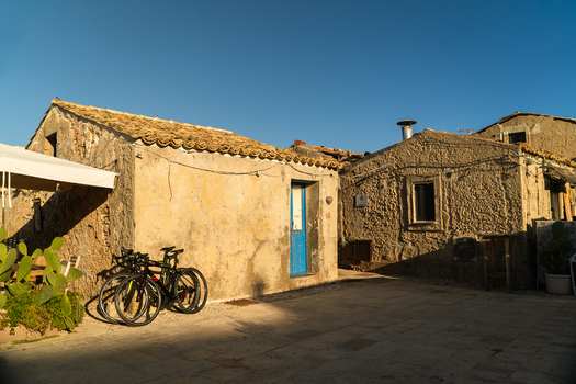 View of rustic stone buildings with a blue door and bicycles parked in a sunlit courtyard under a clear sky in Marsaskala Southern Region Malta. View of rustic stone buildings with a blue door and bicycles parked in a sunlit courtyard under a clear sky in Marsaskala Southern Region Malta.