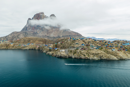 Aerial view of colorful houses in Uummannaq nestled on rocky terrain beneath the iconic Uummannaq mountain with a boat crossing the calm fjord waters in Uummannaq Kitaa Greenland. Aerial view of colorful houses in Uummannaq nestled on rocky terrain beneath the iconic Uummannaq mountain with a boat crossing the calm fjord waters in Uummannaq Kitaa Greenland.