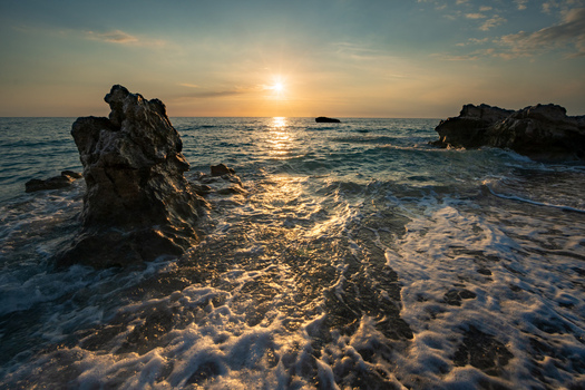 View of golden sunlight reflecting off the shimmering waves crashing around dark jagged rocks at the coast Kathisma Lefkada Greece. View of golden sunlight reflecting off the shimmering waves crashing around dark jagged rocks at the coast Kathisma Lefkada Greece.