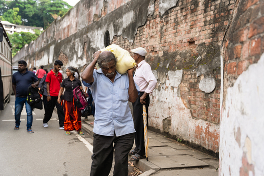 Sri Lanka 20 February 2026 View of a man carrying a heavy yellow sack on his shoulder while walking past a weathered brick wall on a busy street with people and a bus.