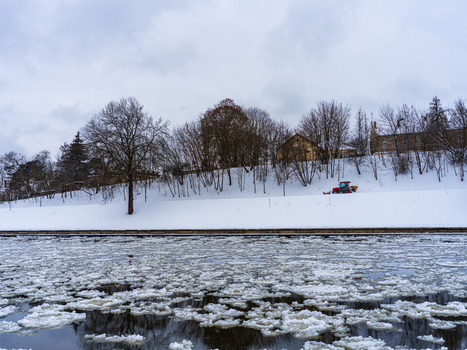 View of the ice covered Neris River with snow covered banks bare trees and a small tractor clearing snow in Vilnius Vilnius County Lithuania. View of the ice covered Neris River with snow covered banks bare trees and a small tractor clearing snow in Vilnius Vilnius County Lithuania.