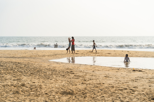 Sri Lanka 16 January 2026 View of children playing on a sandy beach with a woman helping a child do a handstand near crashing waves and a shallow pool of water.