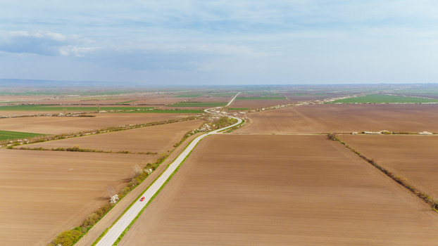Aerial view of a lone red car traveling down a straight road bisecting vast fields of earthy browns and greens under a soft sky Sremska Mitrovica Vojvodina Serbia. Aerial view of a lone red car traveling down a straight road bisecting vast fields of earthy browns and greens under a soft sky Sremska Mitrovica Vojvodina Serbia.