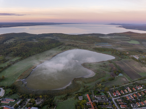 Aerial view of the Inner Lake in Tihany surrounded by rolling hills green fields and residential houses with Lake Balaton in the background under a soft sunrise sky. Tihany Hungary. Aerial view of the Inner Lake in Tihany surrounded by rolling hills green fields and residential houses with Lake Balaton in the background under a soft sunrise sky. Tihany Hungary.