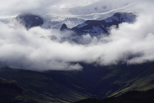 View of Skaftafell mountain range with snow capped peaks partially obscured by low hanging clouds and lush green slopes in Skaftafell Sveitarfelagid Hornaf Iceland. View of Skaftafell mountain range with snow capped peaks partially obscured by low hanging clouds and lush green slopes in Skaftafell Sveitarfelagid Hornaf Iceland.
