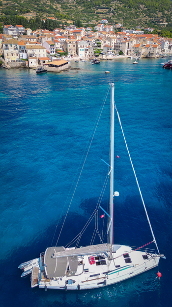 Aerial view of a white sailboat anchored in turquoise waters with the historic coastal town and lush green hills in Komiza Split Dalmatia County Croatia.