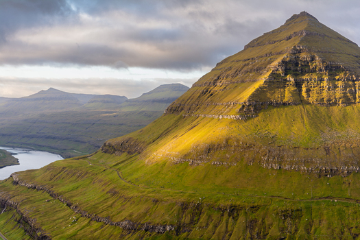 View of a lush green mountain peak illuminated by golden sunlight under a cloudy sky in the Faroe Islands. View of a lush green mountain peak illuminated by golden sunlight under a cloudy sky in the Faroe Islands.