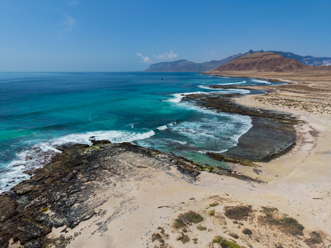 Aerial view of turquoise waters meet rocky shores under a clear sky contrasting with the arid landscape stretching towards distant mountains Erissel Socotra Yemen.