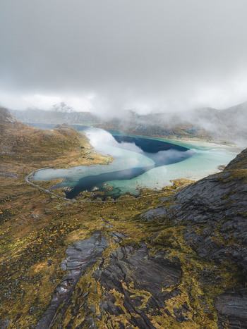 Aerial view of the dramatic Lofoten landscape featuring turquoise fjord waters rugged mountain slopes and a coastal road under a cloudy sky in Leknes Nordland Norway. Aerial view of the dramatic Lofoten landscape featuring turquoise fjord waters rugged mountain slopes and a coastal road under a cloudy sky in Leknes Nordland Norway.