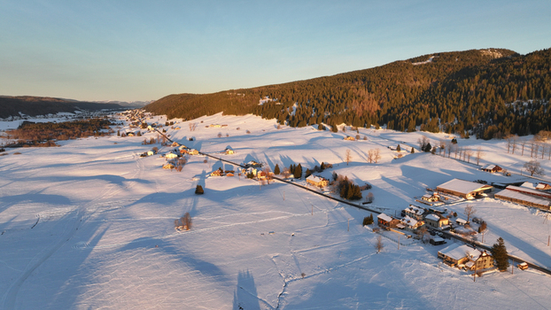 Aerial view of a snow covered landscape with scattered houses nestled against a backdrop of dense forest and undulating hills Les Rousses Bourgogne Franche Comté France. Aerial view of a snow covered landscape with scattered houses nestled against a backdrop of dense forest and undulating hills Les Rousses Bourgogne Franche Comté France.