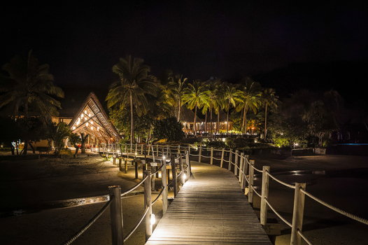 View of the illuminated wooden boardwalk leading to the Likuliku Lagoon Resort at night surrounded by palm trees and tropical foliage in Yaro Nadroga Navosa Fiji.