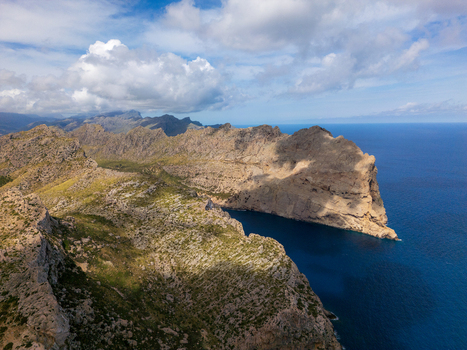 Aerial view of Cap de Formentor featuring rugged limestone cliffs and the deep blue Mediterranean sea under a cloudy sky in Balearic Islands Spain.