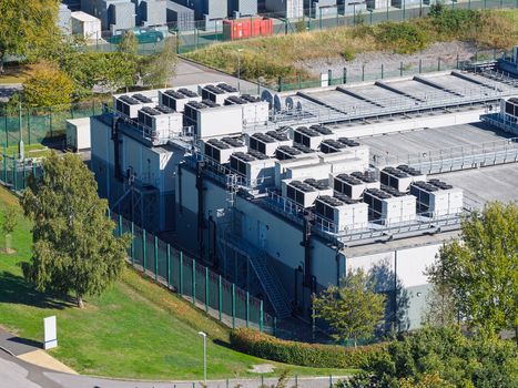 Aerial view of a data center facility with numerous cooling units on the roof surrounded by a green security fence and trees in the United Kingdom.