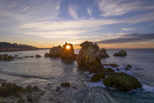 Aerial view of Praia dos Arrifes at sunset with sun shining through a natural rock arch in the ocean calm waves and golden hour light over rocky coastline in Faro District Portugal. Aerial view of Praia dos Arrifes at sunset with sun shining through a natural rock arch in the ocean calm waves and golden hour light over rocky coastline in Faro District Portugal.