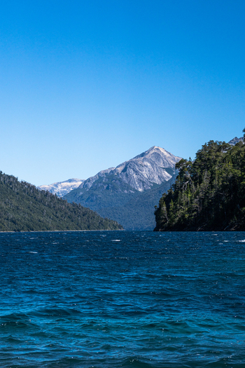 View of the deep blue waters of Nahuel Huapi Lake surrounded by lush forests and snow capped mountains under a clear sky in San Carlos de Bariloche Rio Negro Argentina.