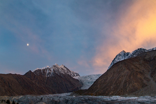 View of a majestic glacier winding through rugged snow capped mountains under a twilight sky with a crescent moon and warm glowing clouds Gilgit Baltistan Pakistan. View of a majestic glacier winding through rugged snow capped mountains under a twilight sky with a crescent moon and warm glowing clouds Gilgit Baltistan Pakistan.