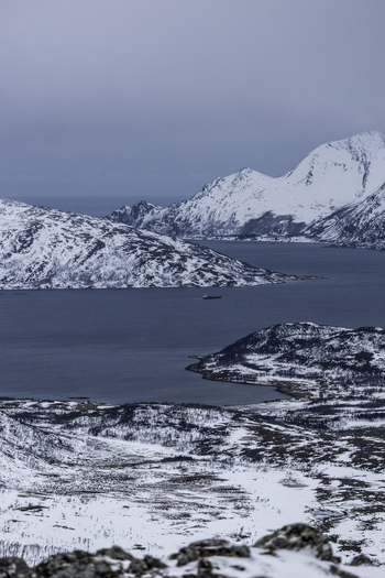 View of snow covered mountains overlooking a deep blue fjord with a distant ship under a moody winter sky Kvaloya Troms Norway.