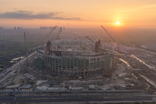 Aerial view of a large stadium under construction with multiple cranes and steel structures at sunset in Vietnam. Aerial view of a large stadium under construction with multiple cranes and steel structures at sunset in Vietnam.