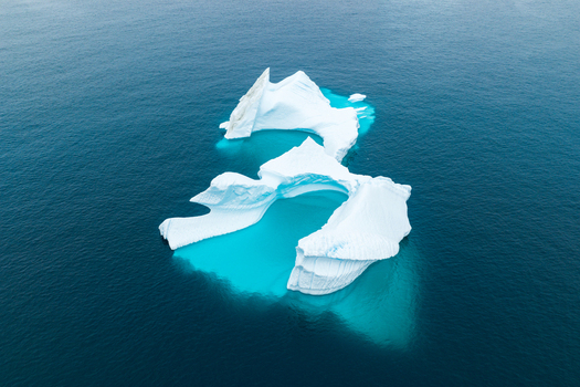 Aerial view of a large jagged white iceberg floating in the deep blue waters of the Arctic Ocean showcasing its intricate ice formations and turquoise submerged base in Greenland.