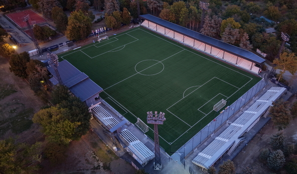 Aerial view of Mladost Sports Center featuring a green artificial turf football pitch surrounded by spectator stands floodlight towers and surrounding trees in Pancevo Serbia. Aerial view of Mladost Sports Center featuring a green artificial turf football pitch surrounded by spectator stands floodlight towers and surrounding trees in Pancevo Serbia.