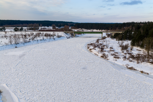 Aerial view of a frozen river covered in ice and snow with a green bridge crossing over it and dense pine forests on the banks in Lithuania. Aerial view of a frozen river covered in ice and snow with a green bridge crossing over it and dense pine forests on the banks in Lithuania.