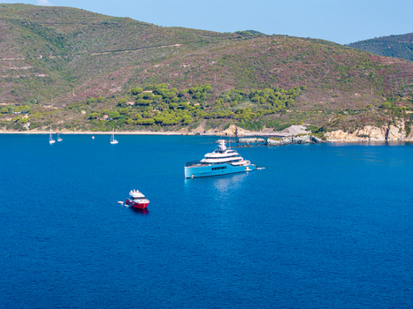 Aerial view of a large luxury yacht and a smaller red motorboat anchored in the clear blue waters of Lacona Bay surrounded by the green hills of Elba island in Lacona Tuscany Italy.
